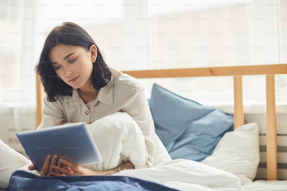 Retrato da mulher adulta moderna usando tablet digital e navegando na internet enquanto se senta na cama confortável pela manhã, espaço de cópia