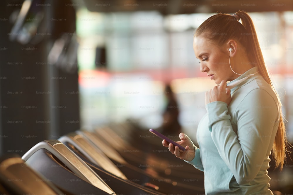 Side view portrait of sportive young woman setting up workout playlist while standing on treadmill in gym, copy space
