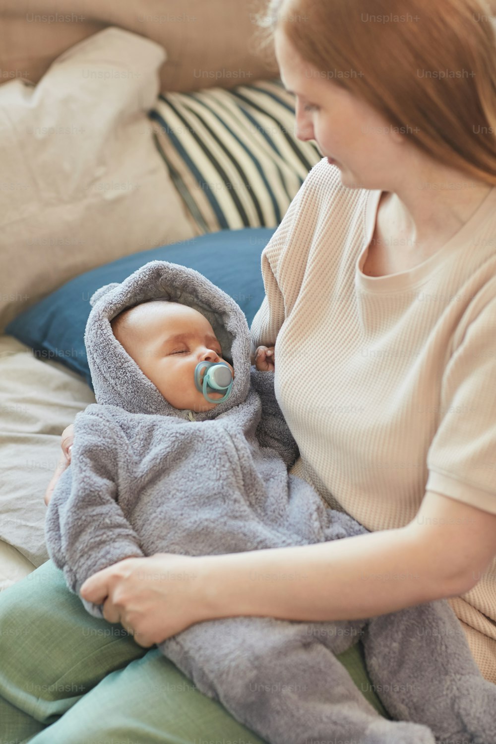 Vertical high angle portrait of young caring mother holding cute sleeping baby in arms while sitting on bed in home interior