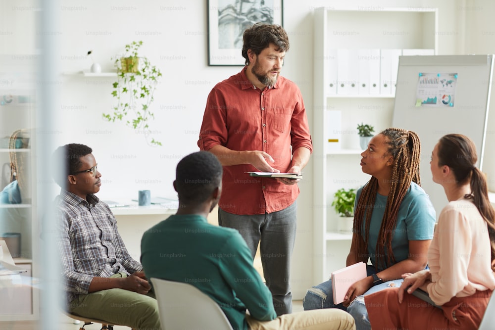 Multi-ethnic group of people sitting in circle while discussing business project in office, focus on smiling beaqrded manager talking to colleagues, copy space