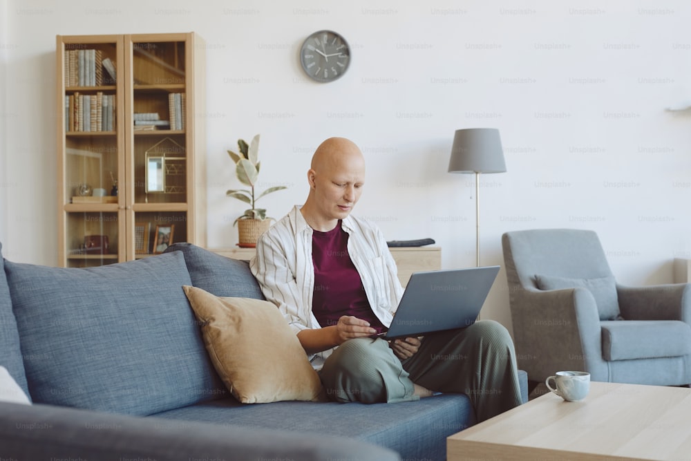 Retrato de una mujer adulta calva usando una computadora portátil mientras está sentada en el sofá en el interior de la casa moderna, la alopecia y la conciencia del cáncer, espacio de copia