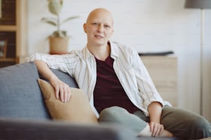 Ritratto di donna adulta calva che guarda la macchina fotografica mentre è seduta sul divano in interni domestici moderni, alopecia e consapevolezza del cancro, spazio di copia