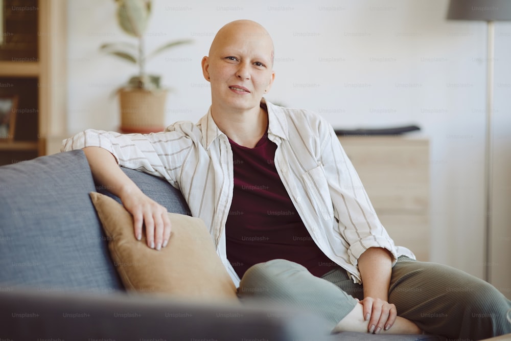 Porträt einer glatzköpfigen erwachsenen Frau, die in die Kamera schaut, während sie auf der Couch in modernem Wohnraum sitzt, Alopezie und Krebsbewusstsein, Kopierraum