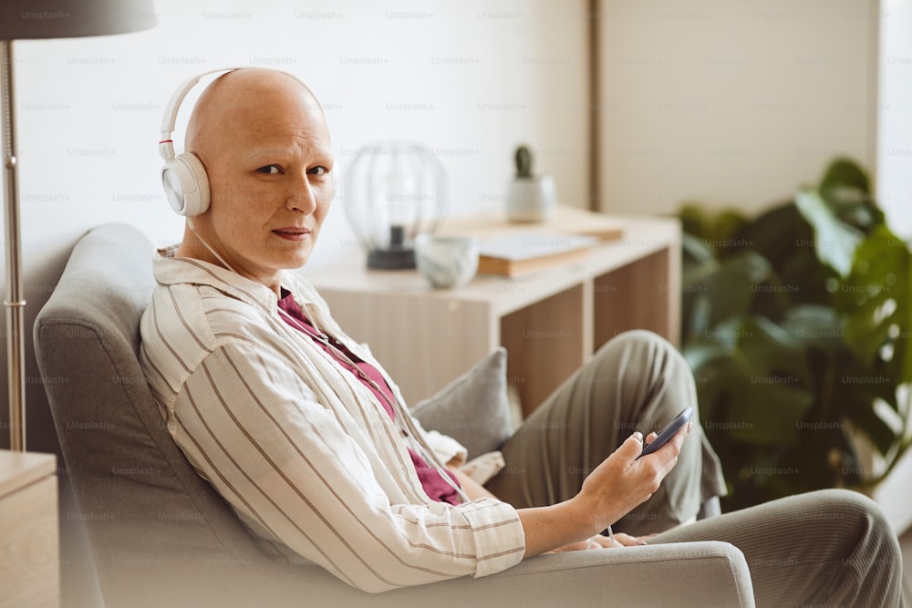 Minimales Seitenansichtsporträt einer glatzköpfigen erwachsenen Frau, die Kopfhörer trägt und in die Kamera schaut, während sie Musik über das Smartphone in gemütlichem Wohnraum, Alopezie und Krebsbewusstsein hört, Kopierraum