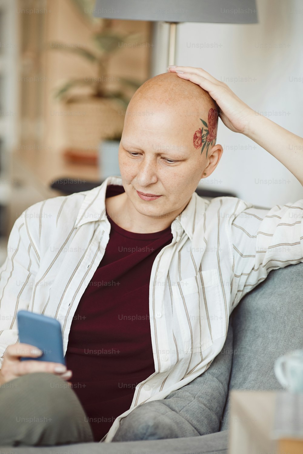Ritratto verticale della donna adulta calva con il tatuaggio della testa usando lo smartphone mentre è seduta in una poltrona accogliente a casa, alopecia e consapevolezza del cancro