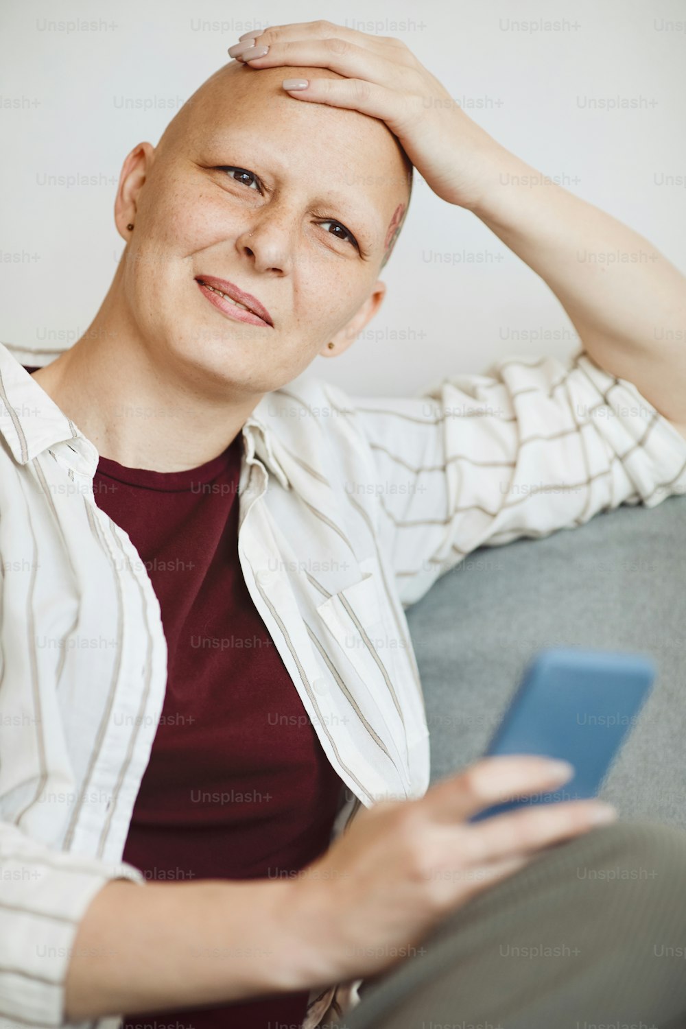 Vertikales Porträt einer lächelnden glatzköpfigen Frau, die sich den Kopf reibt und nachdenklich wegschaut, während sie zu Hause in einem gemütlichen Sessel sitzt, Bewusstsein für Alopezie und Krebs