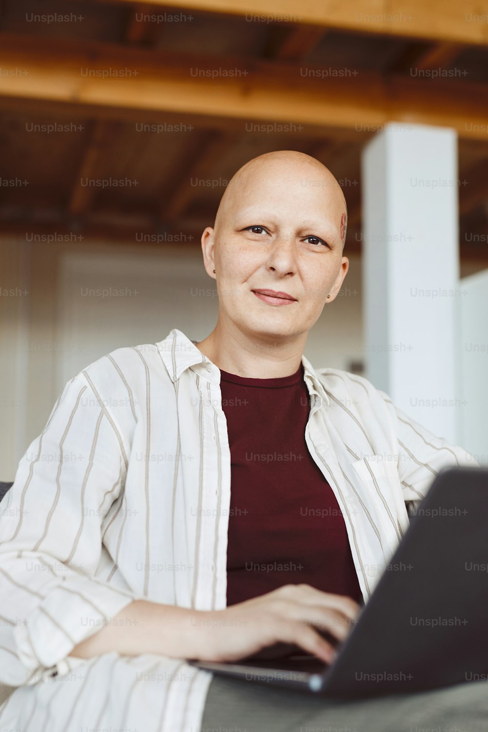 Vertikales Porträt einer glatzköpfigen erwachsenen Frau, die in die Kamera lächelt, während sie einen Laptop hält und zu Hause in modernem Interieur, Alopezie und Krebsbewusstsein arbeitet