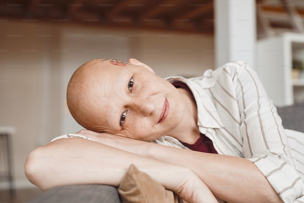 Retrato de close up mínimo de mulher adulta careca sorrindo para a câmera enquanto descansa no sofá no interior da casa em tons quentes, alopecia e consciência do câncer, espaço de cópia