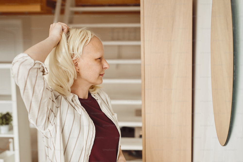 Retrato de vista lateral de tom quente da mulher careca tirando a peruca enquanto está de pé pelo espelho no interior da casa, alopecia e consciência do câncer, espaço de cópia