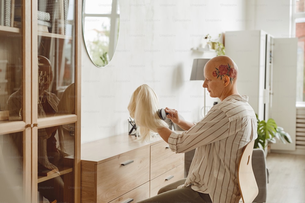Seitenansichtsporträt einer modernen kahlköpfigen Frau, die Perücke bürstet, während sie am Spiegel im Wohnraum sitzt, Alopezie und Krebsbewusstsein, Kopierraum