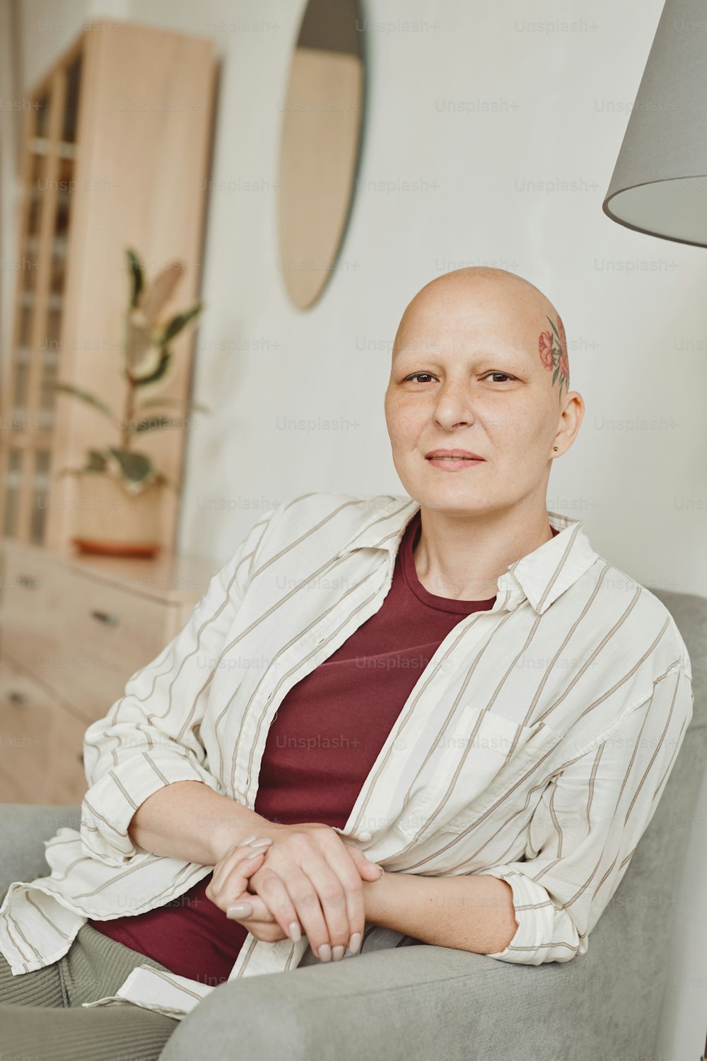 Vertical warm-toned portrait of confident bald woman with head tattoo looking at camera while sitting on couch in home interior, alopecia and cancer awareness