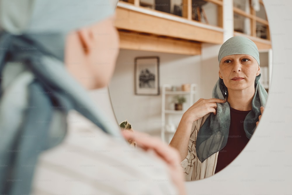 Warmes Porträt einer modernen glatzköpfigen Frau, die ein Kopftuch aufsetzt, während sie in den Spiegel schaut, der im Wohnraum steht, Alopezie und Krebsbewusstsein, Kopierraum
