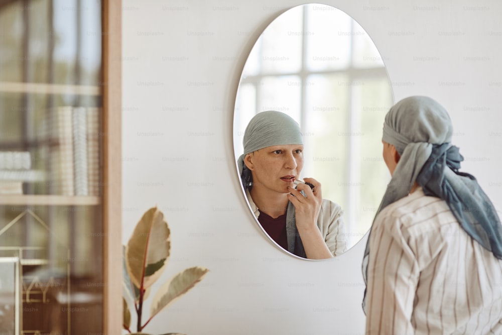 성숙한 대머리 여성이 화장과 립스틱을 바르고 집에서 거울을 보며 아름다움, 탈모증, 암에 대한 인식을 받아들이고 공간을 복사하는 미니멀한 뒷모습