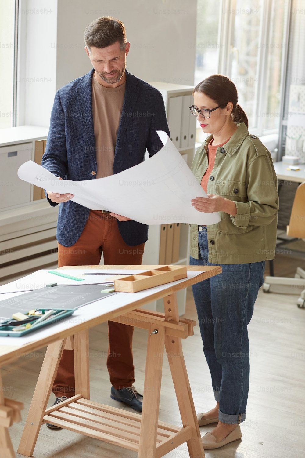 Vertical full length portrait of two architects holding blueprints and discussing work while standing by drawing desk in office
