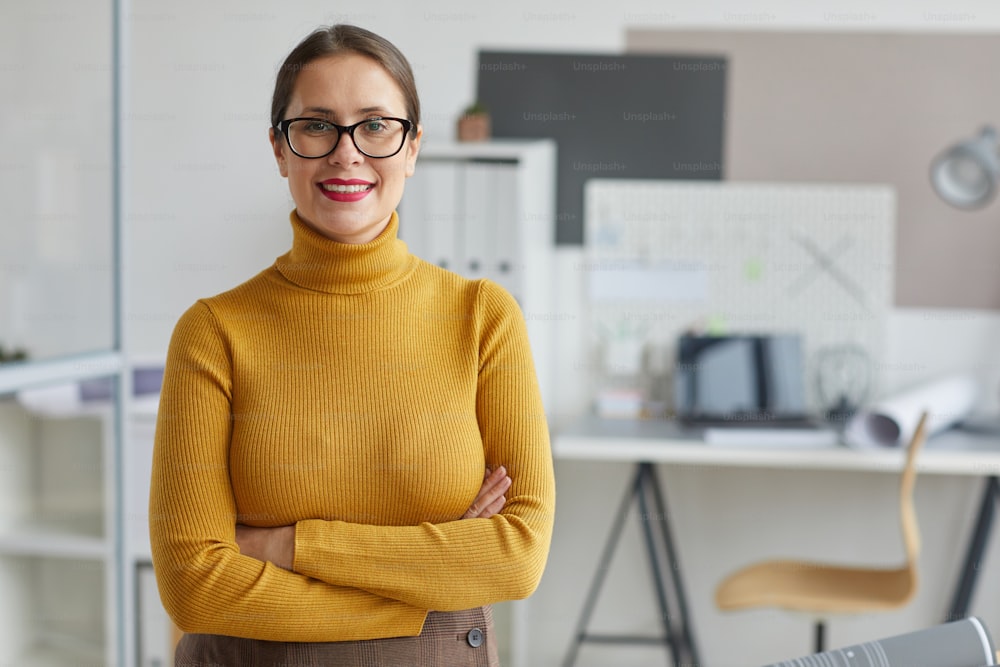 Waist up portrait of smiling female architect standing with arms crossed and looking at camera while posing at workplace in office, copy space