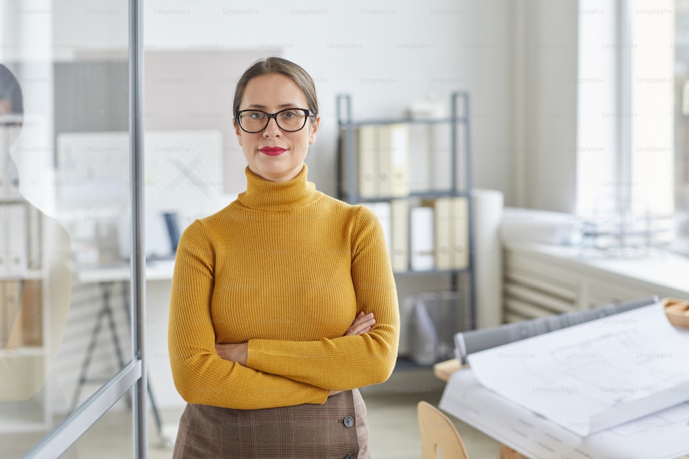 Waist up portrait of confident female architect looking at camera and smiling while standing with arms crossed by drawing desk in office, copy space