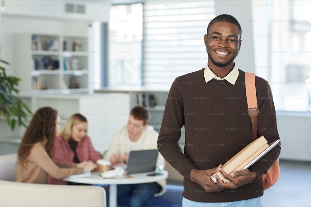 Waist up portrait of smiling African teenage boy looking at camera and smiling while standing in college library with people working in background, copy space