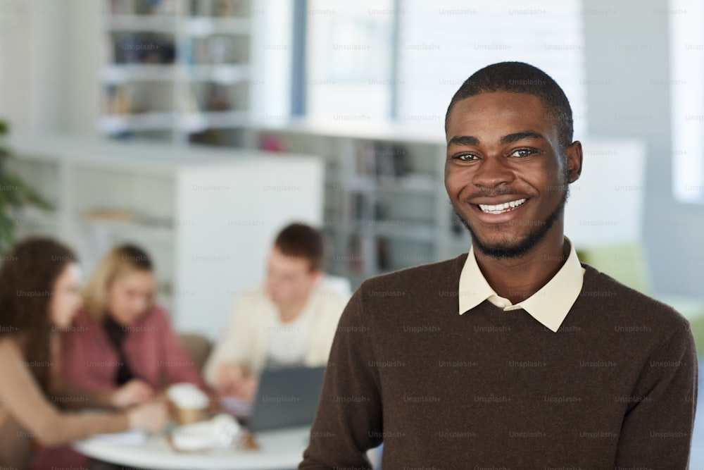 Nahaufnahme des Porträts eines lächelnden afrikanischen Teenagers, der in die Kamera schaut und lächelt, während er in der College-Bibliothek steht, mit Leuten, die im Hintergrund arbeiten, Kopierraum