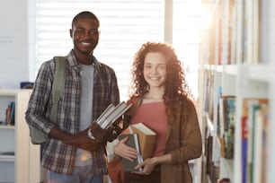 Retrato de cintura para arriba de un hombre afroamericano parado en la biblioteca de la universidad con una mujer joven, sosteniendo libros y sonriendo a la cámara.
