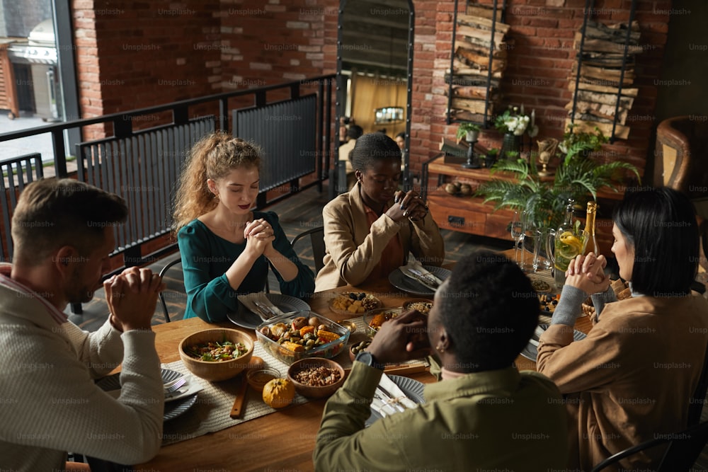 Retrato de alto ángulo de un grupo multiétnico de jóvenes elegantes rezando con los ojos cerrados mientras están sentados a la mesa durante la celebración del Día de Acción de Gracias, espacio de copia