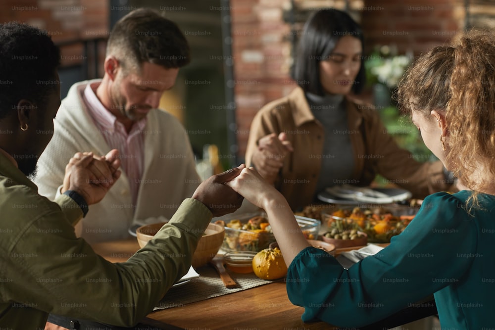Close up portrait of multi-ethnic group of elegant young people praying and holding hands while sitting at dinner table during Thanksgiving celebration, copy space