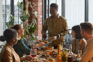 Portrait of smiling African-American man serving food while hosting dinner party with friends and family at home, copy space