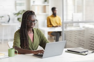 Ritratto di donna afroamericana contemporanea che utilizza il computer portatile mentre lavora alla scrivania in interni bianchi dell'ufficio, spazio di copia