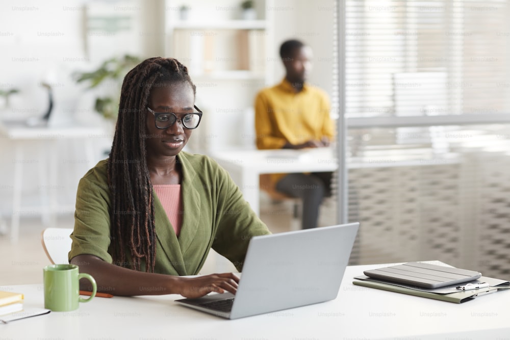 현대 아프리카계 미국인 여성의 초상화는 하얀 사무실 내부의 책상에서 일하는 동안 노트북을 사용하며, 공간을 복사한다