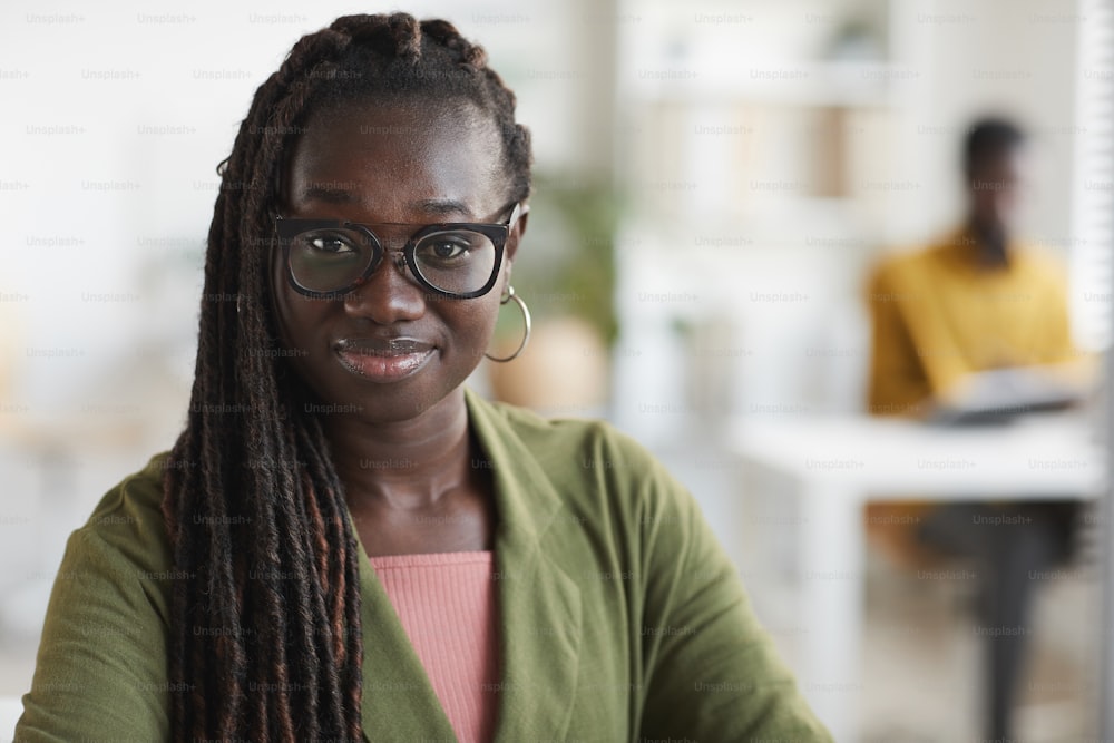 Retrato de cabeza y hombros de una joven afroamericana mirando a la cámara y sonriendo mientras disfruta del trabajo en la oficina, espacio de copia