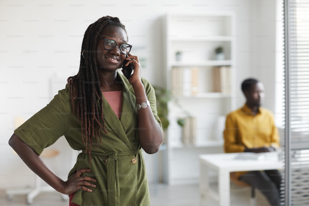 Waist up portrait of stylish young African woman speaking by phone in office and smiling at camera, copy space