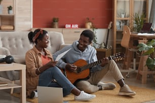 Ritratto a figura intera di due giovani musicisti afroamericani che suonano la chitarra e scrivono musica insieme mentre sono seduti sul pavimento in studio di registrazione, spazio copia