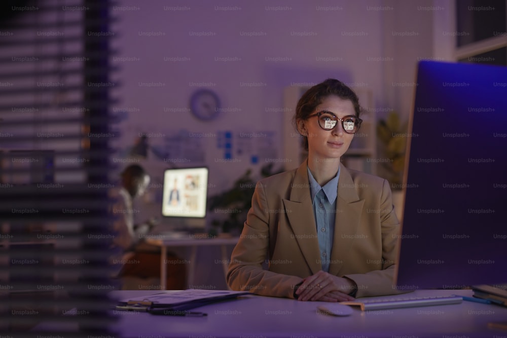 眼鏡をかけた美しい若い女性が夜遅くにオフィスの机に座ってコンピューターのモニターで何かを見ている