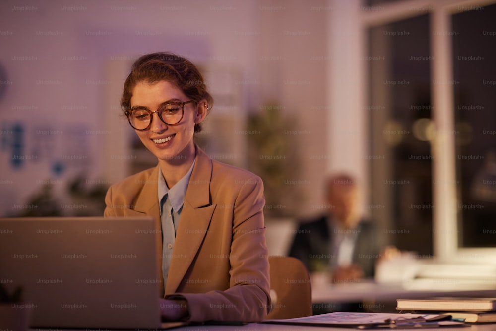 Portrait of joyful young woman wearing eyeglasses sitting at desk in office using laptop late in evening