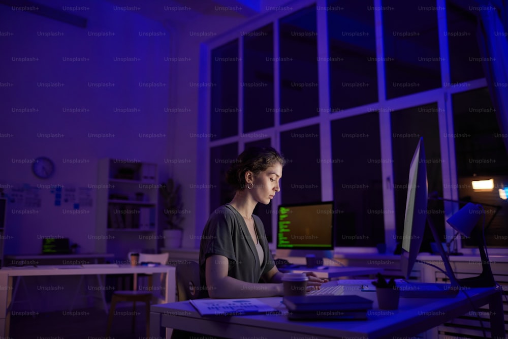 Moderne junge Frau, die spät abends in einem dunklen Büroraum sitzt, Kaffee trinkt und auf einen Computermonitor schaut