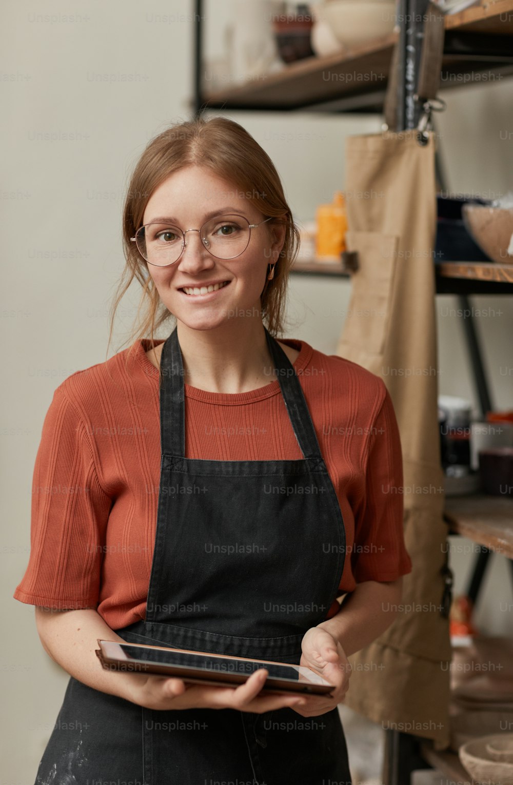 Retrato vertical de cintura para arriba de una joven artesana sosteniendo una tableta y sonriendo a la cámara mientras administra una pequeña empresa en un taller de cerámica