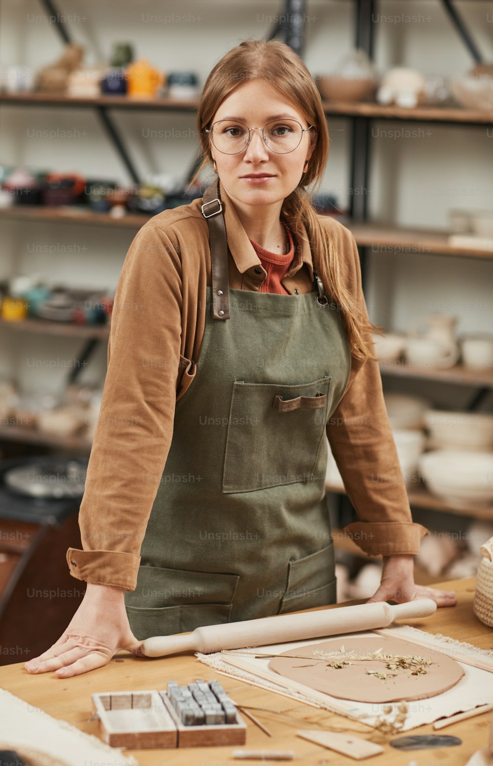 Warm toned vertical portrait of young female artisan looking at camera while making ceramics standing by workstation in pottery workshop