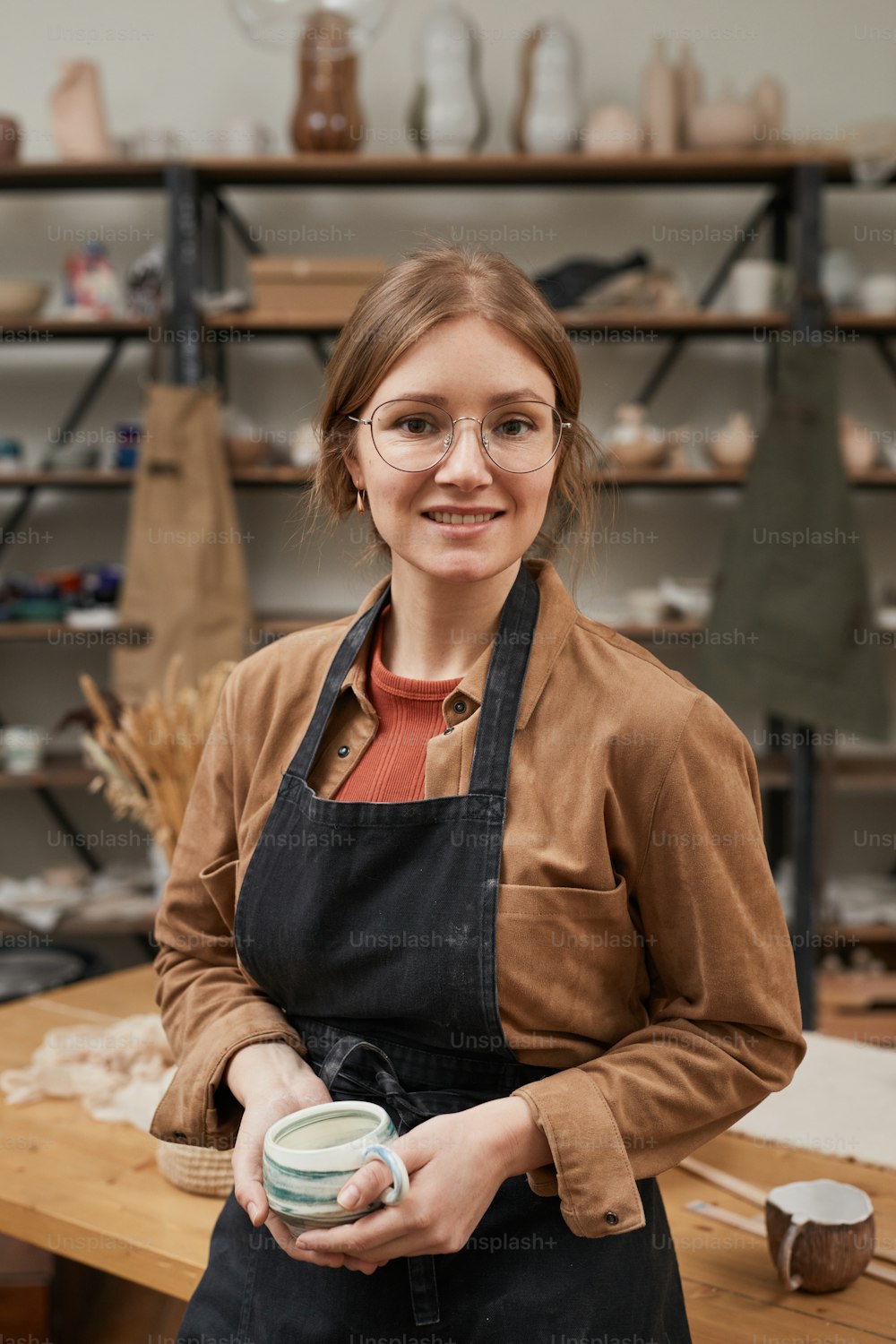 Retrato vertical de cintura para arriba de una joven alfarera mirando a la cámara mientras posa en un taller, pasatiempo y pequeña empresa