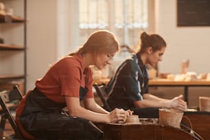 Side view portrait of two young people shaping clay on pottery wheel while making ceramics in workshop lit by sunlight, copy space