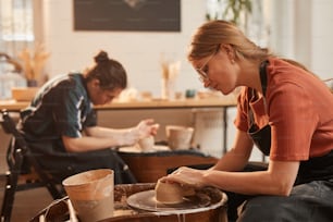 Seitenansichtsporträt einer jungen Frau, die Ton auf einer Töpferscheibe formt, während sie Keramik in einer von Sonnenlicht beleuchteten Werkstatt herstellt, Kopierraum