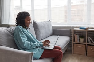 Side view portrait of young African American woman using laptop and smiling while sitting on sofa at home with focus on skin imperfections, copy space
