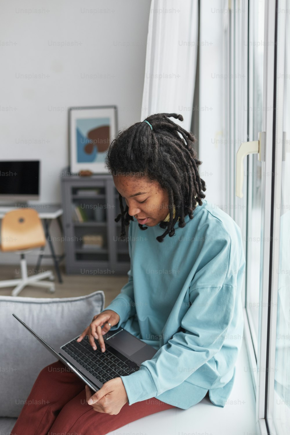 Retrato vertical da mulher afro-americana real usando laptop enquanto sentada na janela em casa com foco nas imperfeições da pele