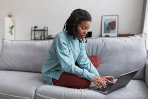 Retrato de corpo inteiro da mulher afro-americana real usando laptop enquanto está sentado no sofá em casa com foco nas imperfeições da pele, espaço de cópia