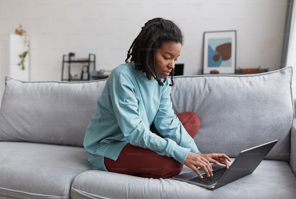 Retrato de cuerpo entero de una mujer afroamericana real usando una computadora portátil mientras está sentada en el sofá de su casa con enfoque en las imperfecciones de la piel, espacio de copia