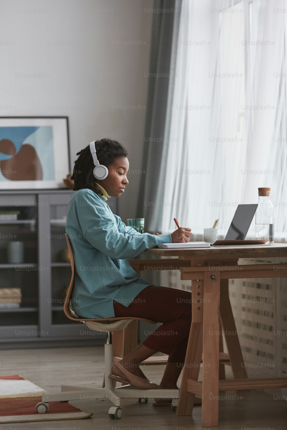 Vista lateral de corpo inteiro em uma jovem mulher afro-americana que estuda em casa ou trabalha na mesa enquanto usa fones de ouvido