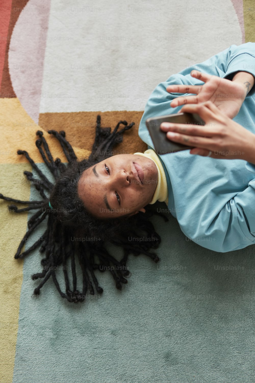 Top view portrait of real African American woman using smartphone or taking selfie while lying on floor on colorful rug, , focus on skin imperfections