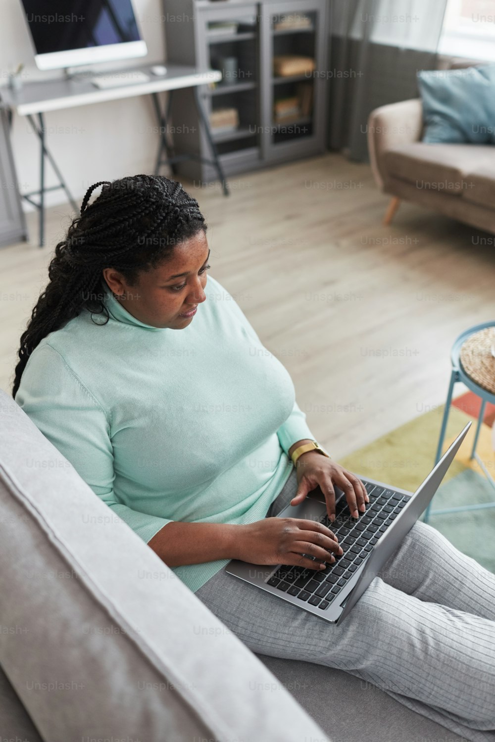 Vertikales Porträt einer kurvigen Afroamerikanerin, die einen Laptop benutzt, während sie die Arbeit von zu Hause aus genießt und sich auf der Couch in einem minimalistischen grauen Innenraum entspannt