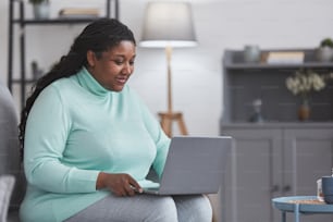 Portrait of curvy African American woman using laptop and smiling while enjoying work from home sitting on couch in minimal grey interior, copy space