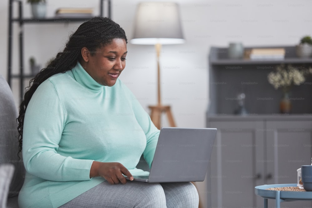 Portrait d’une femme afro-américaine courbée utilisant un ordinateur portable et souriant tout en profitant du travail à domicile assise sur un canapé dans un intérieur gris minimal, espace de copie