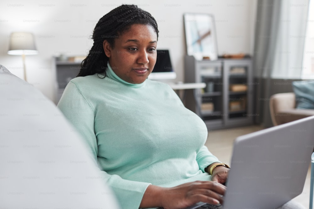 Porträt einer kurvigen Afroamerikanerin, die einen Laptop benutzt, während sie die Arbeit von zu Hause aus genießt und sich auf der Couch in einem modernen, minimalistischen Interieur entspannt