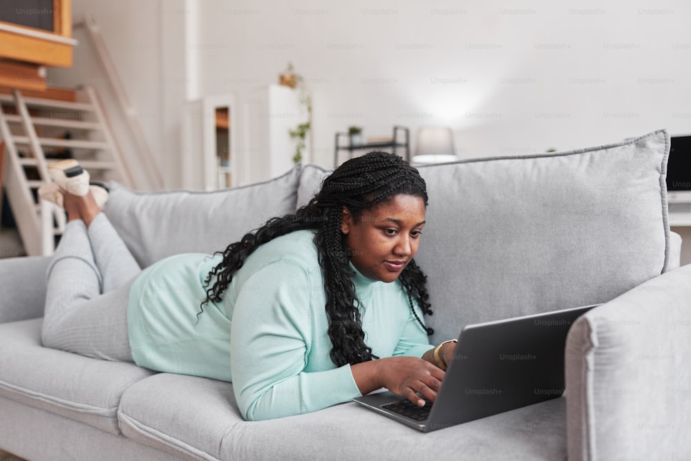 Retrato de cuerpo entero de una mujer afroamericana con curvas usando una computadora portátil mientras está acostada en el sofá y relajándose en casa en un interior minimalista